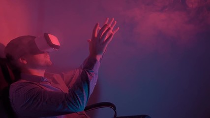 Cute guy in shirt using VR headset interacts with virtual reality. He sits in a smoky dark room with red and blue lighting and waves his hands