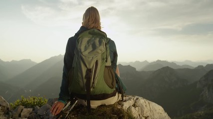 sun flare, close up: unrecognizable female hiker sitting on a rock and observing the breathtaking mo