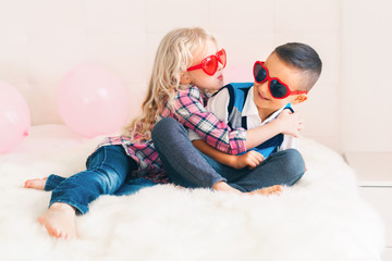 Portrait of two happy white Caucasian cute adorable funny children wearing heart shape glasses. Girl trying to kiss a boy. Love, friendship and fun. Valentine day holiday celebration.