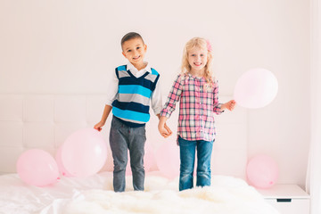 Group portrait of two happy white Caucasian cute adorable funny children standing on bed in bedroom. Boy and girl having fun with pink balloons. Love, friendship and fun. Best friends forever.