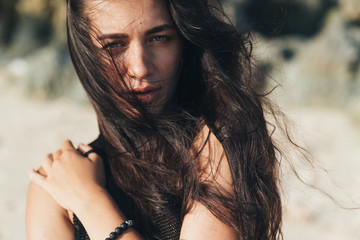 Portrait of sexy brunette model in knitted sweater posing on the sandy beach, big stones and rock on background.