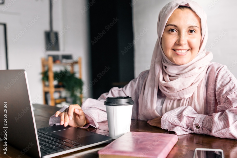 Muslim woman in hijab sitting at a table with a laptop and a glass of ...
