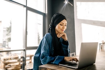 Muslim woman in hijab sitting at a table with a laptop in a cafe. Modern casual Muslim fashion