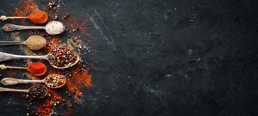 Pepper in a spoon. Colored pepper, sea salt, ground pepper, chili pepper. Top view. On a black background.