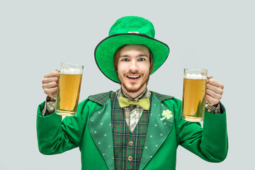 Happy positive young man in green saint patrick suit holding two big mugs of beer and smile. Isolated on grey background.