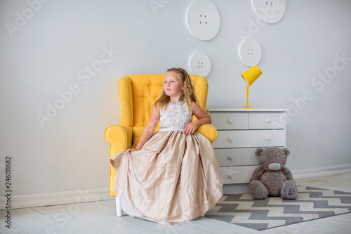 Little Girl In Dress Sitting In An Orange Armchair In Cozy Room