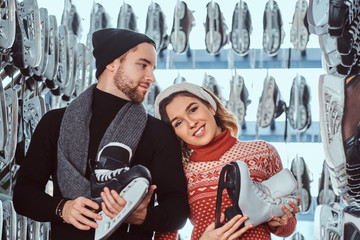 Wall Mural - Young couple wearing warm clothes standing near rack with many pairs of skates, choosing his size, preparing to skate on the ice arena.