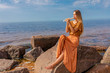 Young girl with long blond hair in long dress playing flute sitting on stone on sea shore