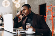 © anatoliycherkas - Portrait of happy african businessman using phone and drinking coffee while working on laptop in a restaurant.