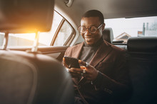 Young Smiling African Man Using Smartphone While Sitting On Backseat In Car. Concept Of Happy Business People Traveling.
