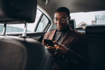 Young smiling african man using smartphone while sitting on backseat in car. Concept of happy business people traveling.