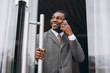 Handsome young african american businessman in classic grey suit holding a smartphone and smiling while leaving the office building