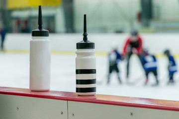 Two white water bottles on the boarder of hockey arena. Blurred Silhouette of the Kids hockey team on the background