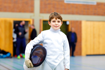 Little kid boy fencing on a fence competition. Child in white fencer uniform with mask and sabre. Active kid training with teacher and children. Healthy sports and leisure.