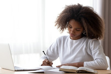 Focused African American Teenage Girl Sit At Table With Laptop Studying With Handbooks, Serious Concentrated Black Teenager Do Homework At Home, Write Down In Notebook, Using Computer And Textbook