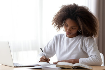 Wall Mural - focused african american teenage girl sit at table with laptop studying with handbooks, serious conc