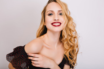 Closeup portrait of young woman, pretty blonde smiling, enjoying, having photoshoot. She has nice soft skin, makeup, long curly hair. Wearing black dress, opened shoulders. Isolated background.