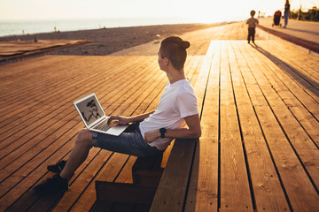 Side view - a slim young student boy sits near the beach on wooden benches and admires the view while studying with a laptop on a sunny summer day. Concept of study in the summer