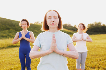 Two generations in family of yoga lovers charming slim woman and two young female daughters are meditating while standing on lawn in a park on a sunny warm summer day. Concept of peace and tranquility