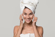 © fizkes - Happy young woman with towel on head apply moisturizing lotion on face look at camera isolated on studio blank background, smiling teen girl use cosmetic cream for healthy skin care, natural beauty