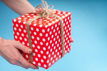 Wall Mural - Hands holding gift box Packed in red polka dot paper on blue background. Holiday card for Valentine's and women's day