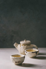 hot green tea in two traditional chinese clay ceramic cup and teapot standing on white marble table.