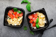 Meal prep. Delicious homemade dinner pasta bows, cherry tomatoes, granulated curd, basil in black plastic containers on a dark background with black forks. Top view
