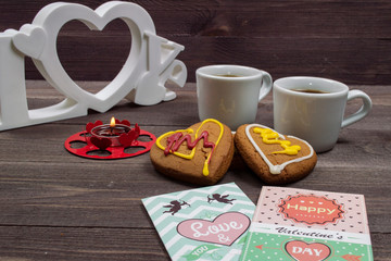 Two white cups of coffee, cookies in the shape of a heart, red candle and a Valentine card on a wooden table on Valentine's Day