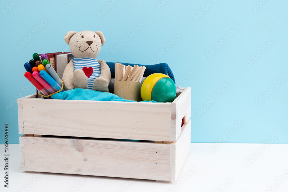 Wooden box with donations: children's toys, stationery and children's clothing. Concept: you can help, they need help. Background, free space for inscription. Red heart on the chest of a teddy bear. - obrazy, fototapety, plakaty 