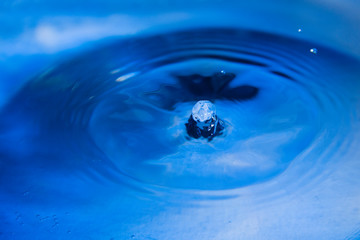 Water droplet falls into the blue water and creates small column close up.