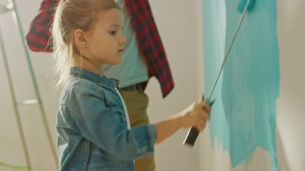 Wall Mural - Adorable Small Girl in Jeans Coat is Painting a Wall. She Paints with Roller that is Covered in Light Blue Paint. Father Paints Together with Daughter. Room Renovations at Home.