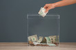 Woman putting money into donation box on table against grey background, closeup