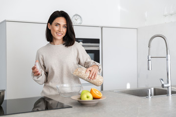 Wall Mural - Image of brunette woman 30s making breakfast with oatmeal and fruits, while standing in modern kitchen at home