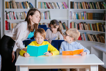 Canvas Print - Education, elementary school, learning and people concept. Three children reading open books in classrom while teacher helping school kids study the subject. Bookshelves on background