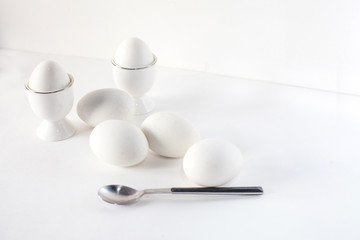 minimalistic composition of two white eggs in a stand and four white eggs lying on a white background with a spoon. copy place for text