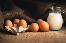 Photo Of Raw Illuminated Eggs  In Kitchen With Jute On Dark Background. Close-up Photography Of Bio Chicken Eggs In Egg Box With Milk In A Ewer Glass.