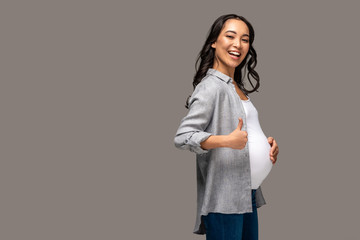 Smiling pregnant asian woman touching belly and showing thumb up isolated on grey