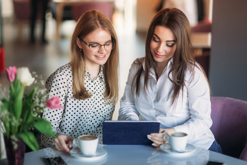 Two girls use a tablet while sitting in a cafe and drinking coffee. Business woman