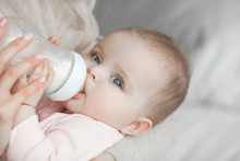 Young Mother Feeding Her Little Cute Baby Daughter With Bottle Of Child Formula. Woman With Her Newborn Baby At Home. Mom Taking Care Of A Child. Alternative To Breast Feeding.