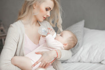 Wall Mural - Young mother feeding her little cute baby daughter with bottle of child formula. Woman with her newborn baby at home. Mom taking care of a child. Alternative to breast feeding.