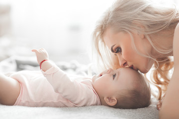 Wall Mural - Young mother taking care of her little baby girl. Beautiful mom and her daughter indoors in the bedroom. Loving family. Attractive mum holding her child.