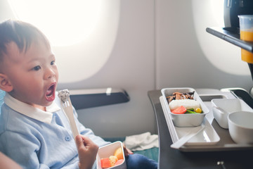 Wall Mural - Cute little Asian 2 -3 years old toddler baby boy child wearing blue sweater eating food during flight on airplane. Flying with children, Happy air travel with kids & little traveler concept