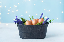 Easter Eggs In An Old Bowl, Grass, Flowers On A Blue Background With Bokeh. Easter And Spring Concept
