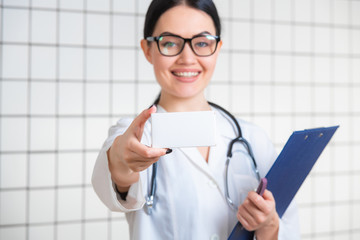 A friendly cute girl with dark hair and glasses, wearing a lab coat, reads carefully the information on the pack in a modern pharmacy.