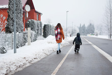 Teen Girl And Little Brother Boy Walking On Pathway Of Street Along Red Brick House In Snowy Frosty Cold Winter Day Back View, Trees Covered With Frost. Family And Children Outdoor Activities Concept.