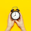 © olgaarkhipenko - Flat lay top view male hand holding black vintage alarm clock on bright yellow color background with copy space. Creative Time concept minimal style, morning time to work