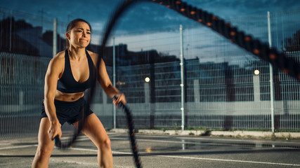 Wall Mural - Beautiful Energetic Fitness Girl Doing Exercises with Battle Ropes. She is Doing a Workout in a Fenced Outdoor Basketball Court. Evening After Rain in a Residential Neighborhood Area.