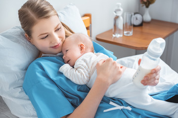 Wall Mural - high angle view of smiling young mother holding baby bottle with milk and lying in bed with adorable baby