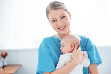 Wall Mural - happy young mother holding newborn baby and smiling at camera in hospital room