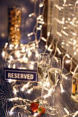 Closeup strawberry, two crystal glasses, bottle champagne sparkling wine, reserved nameplate on wooden table, window, garlands. Concept date evening in restaraunt, 8 march, Valentine's Day, christmas